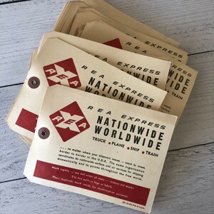 May include: A stack of vintage REA Express shipping tags. The tags are cream-colored with red and black text. The text reads "REA Express Nationwide Worldwide" and includes transportation options: truck, plane, ship, and train.