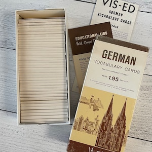 May include: A set of vintage German vocabulary cards in a box. The box and cards are beige and white. The box has the words "GERMAN VOCABULARY CARDS" printed on it, along with an illustration of a cathedral and people. The price is $1.95 per set.