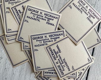 Puede incluir: Un conjunto de tarjetas de recetas antiguas. Cada tarjeta presenta el nombre "GEORGE H. HECKMAN, Prescription Druggist" y los detalles de la dirección en una fuente azul oscuro. Las tarjetas son rectangulares con un borde azul oscuro y la palabra "Dr." en la esquina.