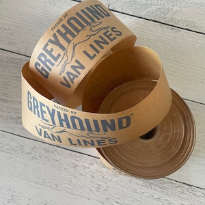 May include: Brown paper tape rolls with the text "PACKED BY GREYHOUND VAN LINES" in dark blue. The Greyhound logo is also visible. The tape is unrolled in a spiral shape, with a roll of tape in the background.