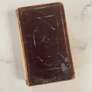 Op de afbeelding: Een vintage, rechthoekig boek met een donkerbruine leren kaft. De kaft heeft een gedetailleerd reliëfontwerp en vertoont tekenen van slijtage. De randen van het boek zijn lichtbruin.