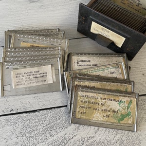 May include: A collection of vintage seed packets and a metal card catalog. The packets, with metal frames, feature labels with text like "Onion to Watermelon" and "Certified Watermelon." The catalog is dark metal, with a label holder.