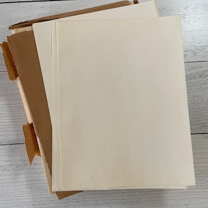 May include: A stack of cream-colored paper sheets next to a brown cardboard box. The paper has a smooth texture and is stacked neatly. The box is open, revealing the edges of the paper. The background is a white wooden surface.