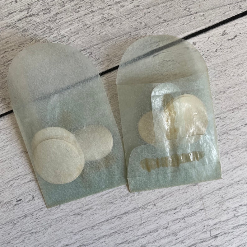 May include: Two small, translucent paper envelopes, each containing several round, light-colored objects. The envelopes are slightly open, revealing the contents. The background is a light-colored, wood-grain surface.