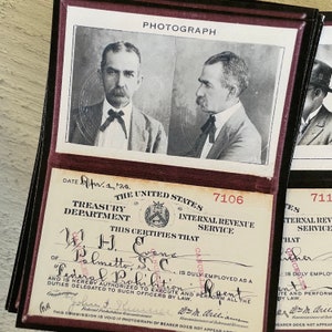 Op de afbeelding: Vintage zwart-witfoto van een man met een snor, in een pak en vlinderdas, met een document van het Amerikaanse ministerie van Financi&euml;n. Het document is van de Internal Revenue Service, met de tekst "Federal Prohibition Agent."