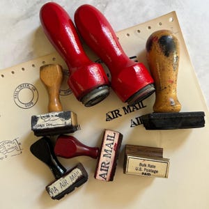 May include: A selection of vintage rubber stamps with wooden handles. The stamps feature the text "AIR MAIL", "VIA AIR MAIL", "Postage Due", and "Bulk Rate U.S. Postage PAID". The handles are red, black, and natural wood tones.