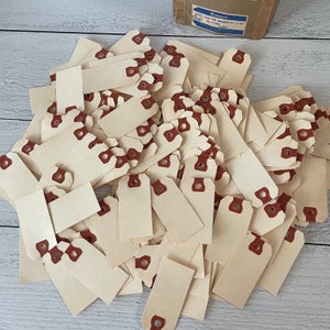 May include: A pile of cream-colored rectangular tags with reinforced brown eyelets. The tags are scattered on a light wood surface. A cardboard box with a blue and white label is in the background.