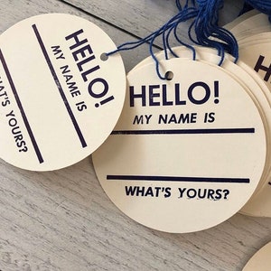 May include: A stack of round, off-white name tags with blue string. Each tag has the words "HELLO! MY NAME IS" printed in dark blue, with a line for writing a name and the phrase "WHAT'S YOURS?"