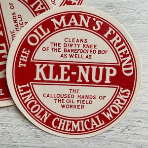 Op de afbeelding: Cirkelvormige vintage papieren advertentie met rode tekst op een crèmekleurige achtergrond. De tekst luidt "The Oil Man's Friend" en "Kle-Nup". De advertentie bevat ook de tekst "Reinigt de vuile knie van de blotevoetenjongen en de eeltige handen van de olieveldarbeider."