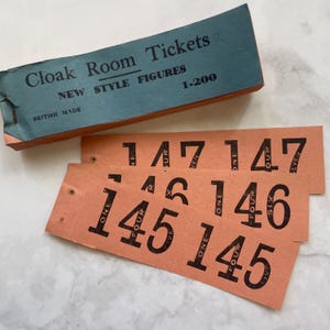 Könnte beinhalten: Ein Satz alter Garderobenmarken. Die pfirsichfarbenen Tickets haben schwarze Zahlen und Text. Die türkisfarbene Box trägt die Aufschrift "Cloak Room Tickets" und "British Made". Die Tickets sind von 1-200 nummeriert.