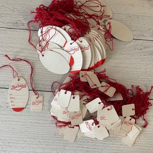 May include: A collection of white True Value price tags with red accents and red string. The tags are in various shapes, including oval and rectangular, and are arranged on a light wood-grain surface.
