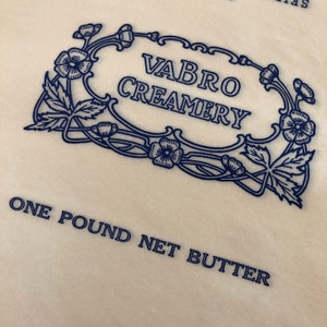 May include: Cream-colored packaging with navy blue text and floral border. The text reads "VABRO CREAMERY" inside a decorative frame. Below, it states "ONE POUND NET BUTTER". The design features stylized flowers and leaves.