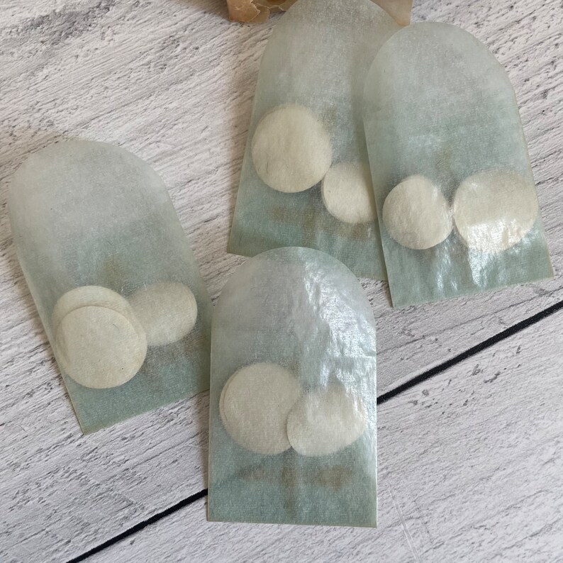 May include: Four small, translucent paper pouches, each containing two or three round, off-white objects. The pouches are arranged on a light-colored, wood-grain surface. The pouches have a rounded top.