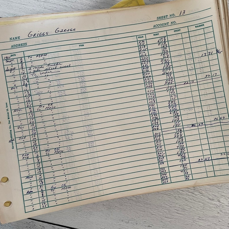 May include: An open ledger book with ruled lines and handwritten entries. The top of the page reads "GRIGGS GARAGE" and "SHEET NO. 13". The entries include dates, items, and financial figures, with a yellow tab visible.