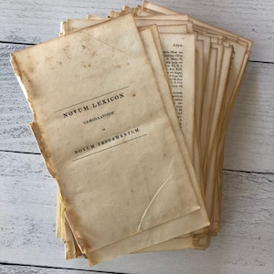 May include: A stack of aged paper pages with the title "NOVUM LEXICON" in black text. The pages are a light tan color with visible wear and tear, suggesting age. The text "GRÆCO-LATINUM" and "NOVUM TESTAMENTUM" are also visible.