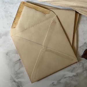 Pode incluir: Uma pilha de envelopes vintage abertos, em tom bege claro. Os envelopes mostram sinais de idade com descoloração e desgaste nas bordas. O papel tem uma aparência ligeiramente texturizada.