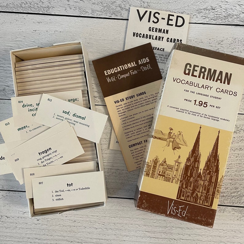 May include: Vintage Vis-Ed German vocabulary cards for language students. The set includes a box of cards with German words and definitions, along with informational pamphlets. The box features a sepia-toned illustration of a cathedral.