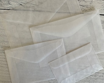 May include: A collection of translucent white envelopes in various sizes. The envelopes are made of a thin, delicate paper, with a flap closure. The envelopes are arranged on a light-colored wooden surface.