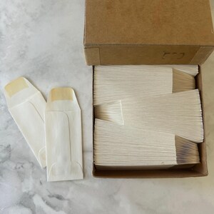 May include: A large cardboard box filled with a stack of small, rectangular, off-white paper envelopes, with two additional identical envelopes placed outside the box to the left, showing their folded flaps and smooth, light-colored paper texture.