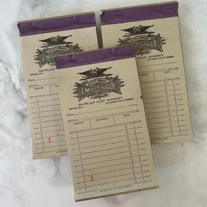 Pode incluir: Três blocos de recibos retangulares de papel com faixas de encadernação superiores roxas. Cada bloco apresenta um logótipo vintage a tinta preta da I. Mattingly Dunlap Hat Agency, com linhas impressas para nome, morada e data.