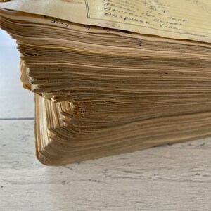 May include: Close-up of a vintage ledger with aged, yellowed pages. The pages are stacked, showing the thickness of the book. Handwritten names and dates are visible on the top page. The book is resting on a white, textured surface.