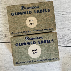 May include: Two vintage Dennison gummed label boxes. The boxes are light green with dark blue text that reads "GUMMED LABELS". Each box has a circular sticker with "1000" and a label number. The boxes are from Framingham, Mass., U.S.A.