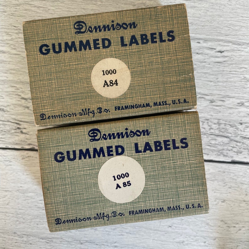May include: Two vintage Dennison gummed label boxes. The boxes are light green with dark blue text that reads "GUMMED LABELS". Each box has a circular sticker with "1000" and a label number. The boxes are from Framingham, Mass., U.S.A.
