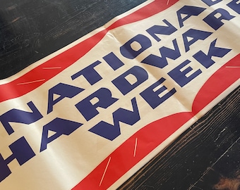 May include: A vintage banner with the text "NATIONAL HARDWARE WEEK" in navy blue letters. The banner has a cream background with red and white accents. The banner is laying on a dark wooden surface.