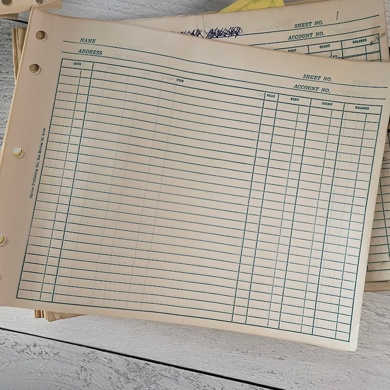 May include: A stack of vintage ledger sheets with green ruled lines and pre-printed headings for accounting records. The sheets have holes for binding and include fields for date, item, debit, credit, and balance. The paper has a slightly aged, off-white color.
