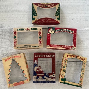 Op de afbeelding: Een verzameling vintage "Shiny Brite" kerstornamentendozen. De dozen hebben feestelijke ontwerpen met afbeeldingen van kerstbomen, de kerstman en sneeuwvlokken. Tekst bevat "American Made Glass Tree Ornaments" en "Star Band Co. New York, N.Y."