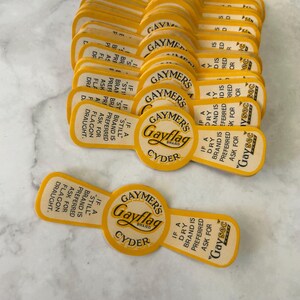 Könnte beinhalten: Eine Sammlung gelb-weißer Vintage-Flaschenetiketten von Gaymer's Gayflag Cyder. Die Etiketten haben eine einzigartige Form und den Text "If a dry brand is preferred ask for Gaysec".