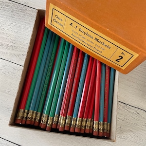 May include: A box of pencils with red, green, and blue painted exteriors. The pencils are labeled "A. J. Bayless Markets" and "YOUR HOME TOWN GROCER". The box is orange and has a label that reads "One Gross" and "No. 2".