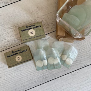 May include: Vintage Dennison gummed labels in boxes, with the text "GUMMED LABELS" and "1000 A85" and "1000 A84". Several small, round, white labels are visible in small translucent bags. A clear plastic container holds more labels.