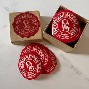Op de afbeelding: Een verzameling rode en witte ronde stickers van The Okemah National Bank, Okemah, Oklahoma, wordt getoond in een kleine kartonnen doos en op een wit oppervlak. De stickers hebben een zegelontwerp met de naam en locatie van de bank.