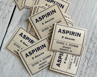 Puede incluir: Una colección de etiquetas de aspirina vintage. Cada etiqueta es cuadrada con fondo crema y texto negro. Las etiquetas dicen "ASPIRIN 5 GRAINS" e incluyen el nombre "GEORGE H. HECKMAN DRUGGIST NAZARETH, PA."