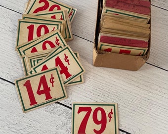 Peut inclure: An assortment of vintage price tags with red numbers and cent symbols, outlined in green. Some tags are scattered, others are in a cardboard box. The tags are printed on aged paper, with the text "MADE IN U.S.A."