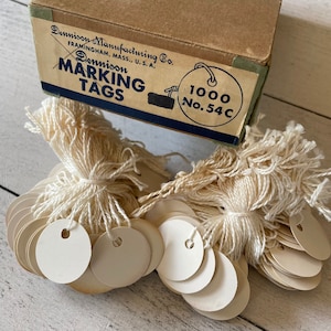 Peut inclure: Une boîte vintage d'étiquettes de marquage Dennison, avec le texte "Dennison Marking Tags" et "1000 No. 54C". Des paquets d'étiquettes rondes de couleur crème sont attachés avec de la ficelle blanc cassé. La boîte est marron avec une étiquette bleue et verte.