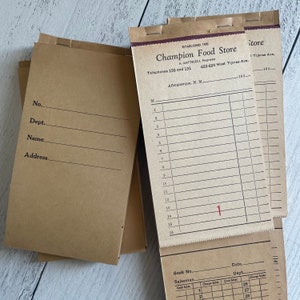 May include: A collection of vintage brown paper notepads. The top notepad displays the text "Champion Food Store" and address details. The notepads have lines for itemizing purchases and other details. The notepads are stapled together at the top.