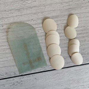 May include: An assortment of communion wafers and a pale green paper tag with a cross design. The wafers are circular and off-white, arranged in two columns and a single row. The tag has a rounded top.