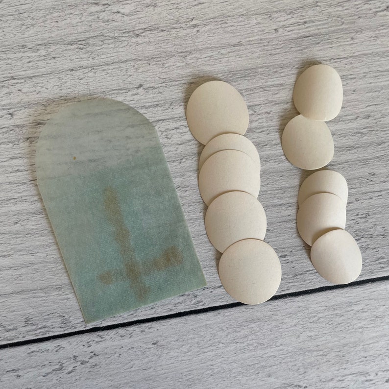 May include: An assortment of communion wafers and a pale green paper tag with a cross design. The wafers are circular and off-white, arranged in two columns and a single row. The tag has a rounded top.