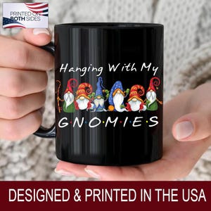 May include: Black ceramic mug featuring the phrase "Hanging With My Gnomies" with a colorful illustration of gnomes. The mug has a black handle and the text "PRINTED ON BOTH SIDES" and "DESIGNED & PRINTED IN THE USA".