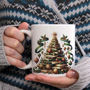 May include: White ceramic mug featuring a Christmas tree design made of books, ornaments, and a star. Green ribbons with the words "Merry Christmas" are wrapped around the tree. The mug has a white handle and is held by a person.