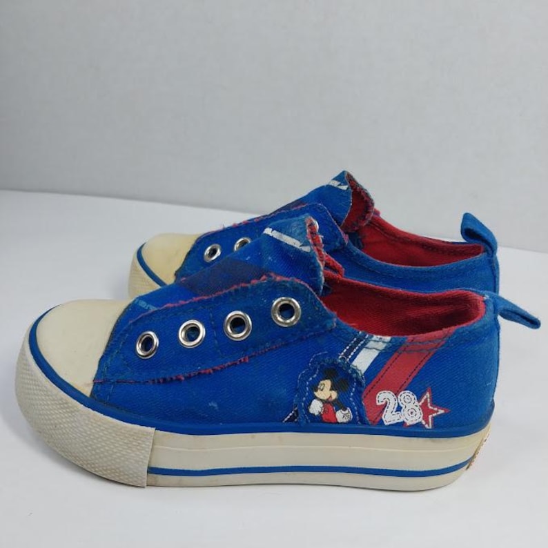 Op de afbeelding: Paar blauwe canvas sneakers met witte neusdoppen en zolen. De schoenen hebben een Mickey Mouse-afbeelding en het nummer 28 aan de zijkant. De binnenvoering is rood.