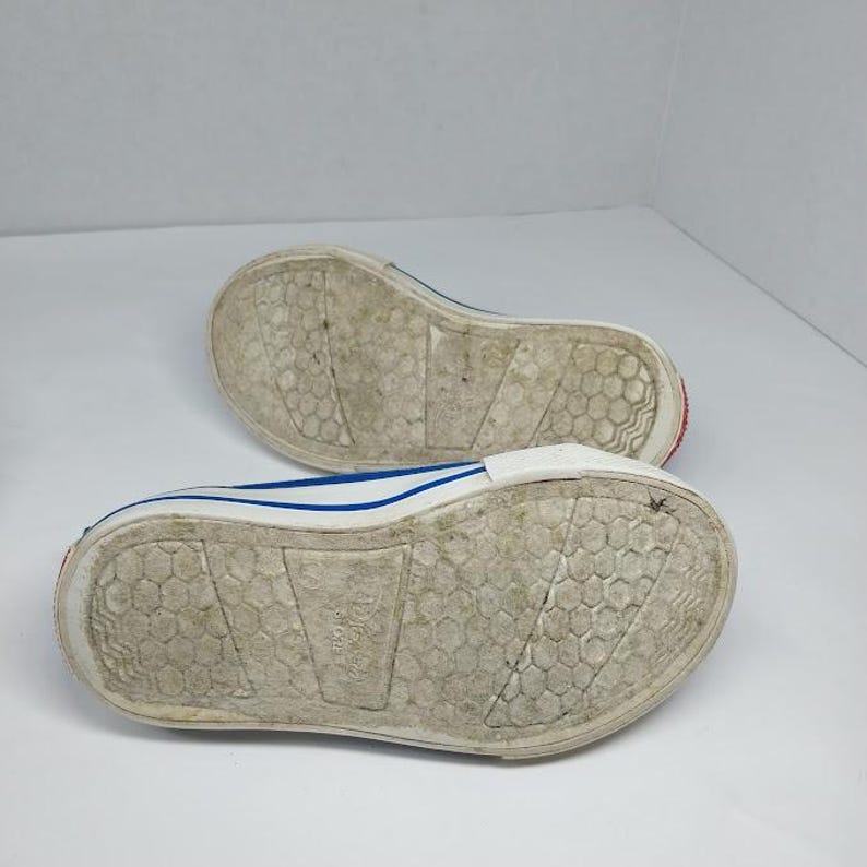 Op de afbeelding: Een paar witte canvas sneakers met blauwe randen en rode accenten op de zolen. De zolen zijn gebroken wit met een textuurpatroon. De schoenen worden van onderaf getoond.