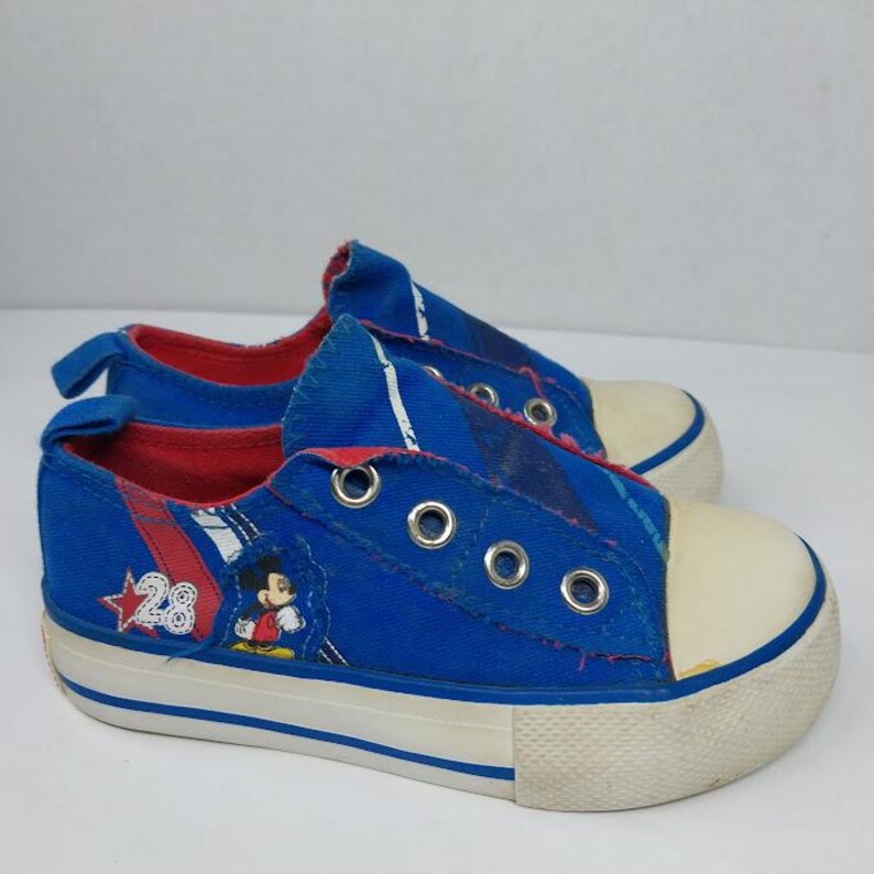 Op de afbeelding: Blauwe canvas kindersneakers met een witte rubberen neus en zool. De sneakers hebben een Mickey Mouse-afbeelding en het nummer 28. De binnenkant is rood.