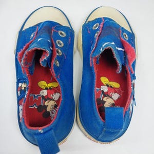 Op de afbeelding: Paar blauwe canvas sneakers met witte rubberen zolen en neus. De binnenkant is rood met een Mickey Mouse-afbeelding. De schoenen hebben metalen vetergaten en een blauwe treklus op de hiel. Het canvas is versleten.