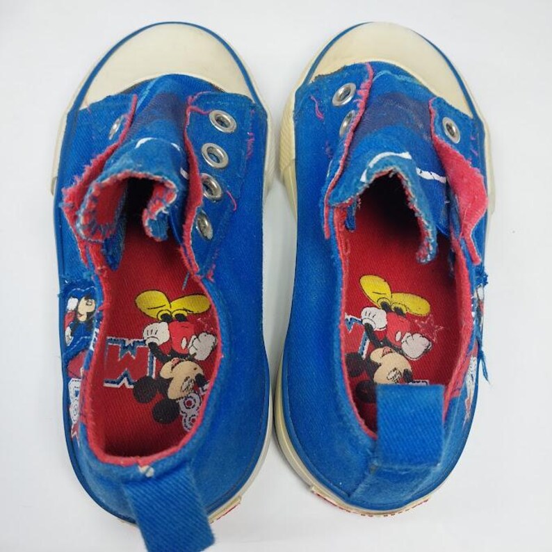 Op de afbeelding: Paar blauwe canvas sneakers met witte rubberen zolen en neus. De binnenkant is rood met een Mickey Mouse-afbeelding. De schoenen hebben metalen vetergaten en een blauwe treklus op de hiel. Het canvas is versleten.