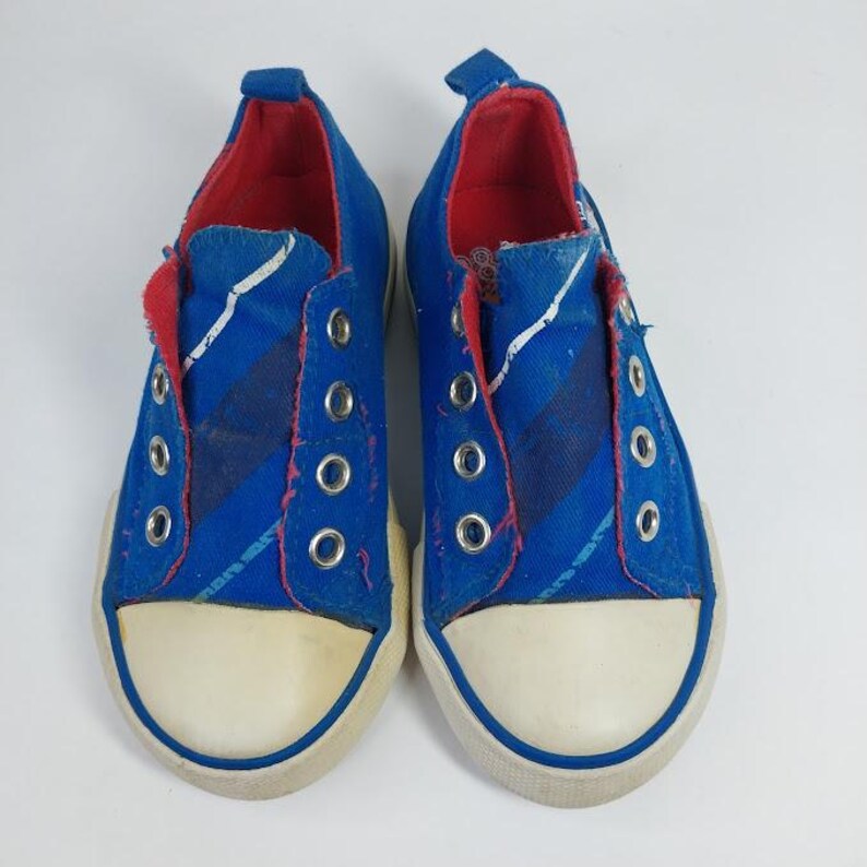 Op de afbeelding: Een paar blauwe canvas sneakers met witte rubberen neuzen en zolen. De schoenen hebben een rode voering en veterogen. Het canvas heeft een gestreept ontwerp in donkerder blauw. De schoenen zijn ontworpen voor kinderen.