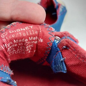 Op de afbeelding: Close-up van de binnenkant van een rode en blauwe schoen. De rode stof heeft een Disney-label met tekst. Blauwe stiksels en accenten zijn zichtbaar. De schoen lijkt van stof te zijn.