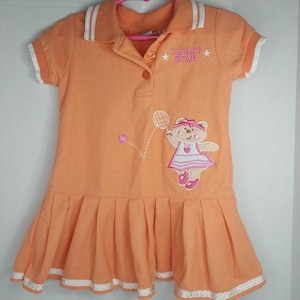 May include: Orange polo dress with white trim on the collar and sleeves. The dress features an embroidered bear playing tennis, with the text "TENNIS STAR". The skirt is pleated.